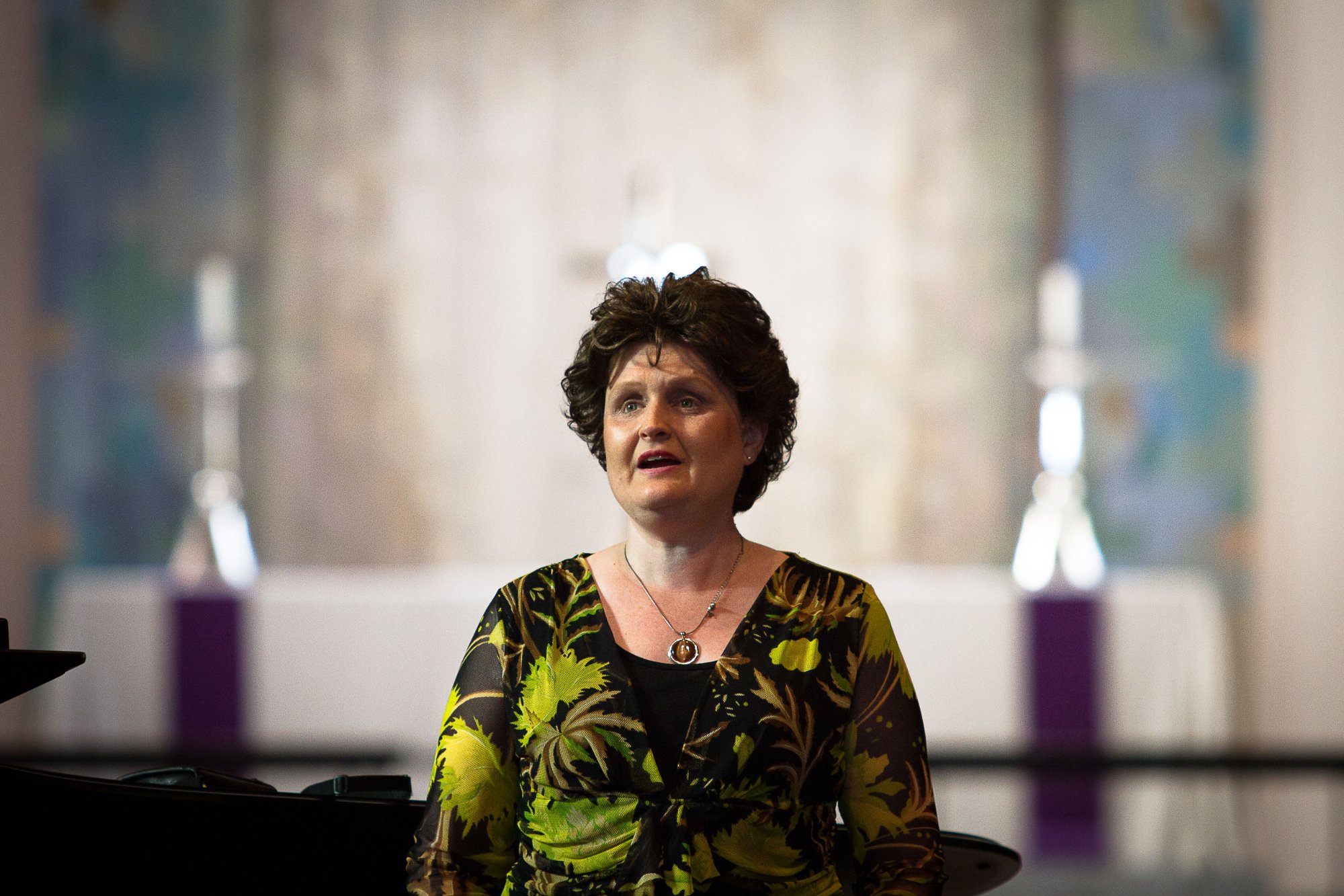 Lisette Wesseling performing in a church, wearing a green and black patterned dress, singing with candles visible in the background